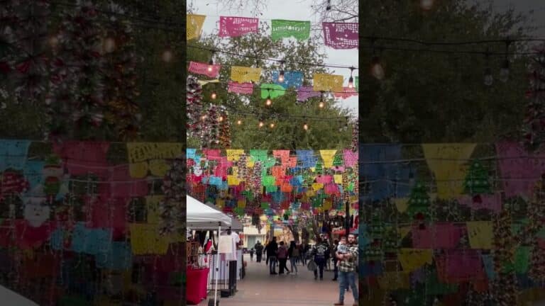 Colorful papel picado banners hang above an outdoor market with people walking among stalls and trees in the background. String lights are also visible overhead.